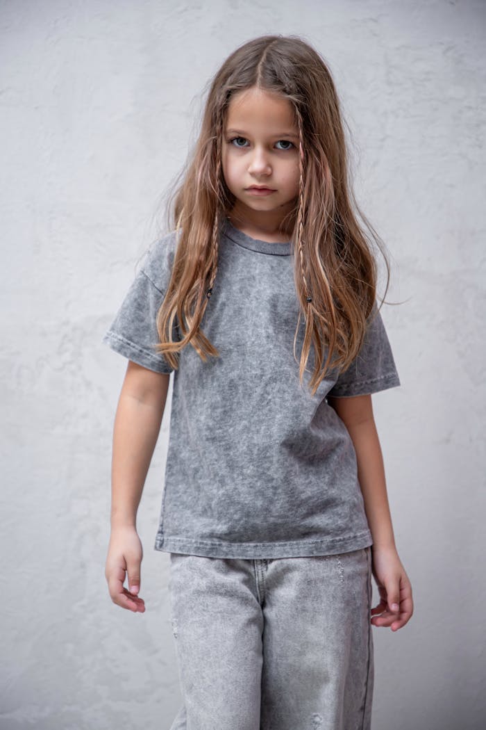 Little girl with long hair wearing gray t-shirt and denim jeans against a neutral background.