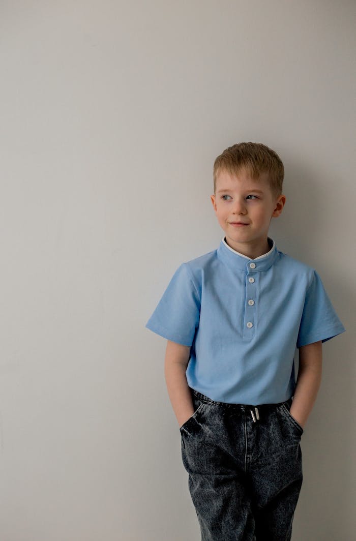 A young boy in a blue shirt and jeans standing against a light wall, looking thoughtful.